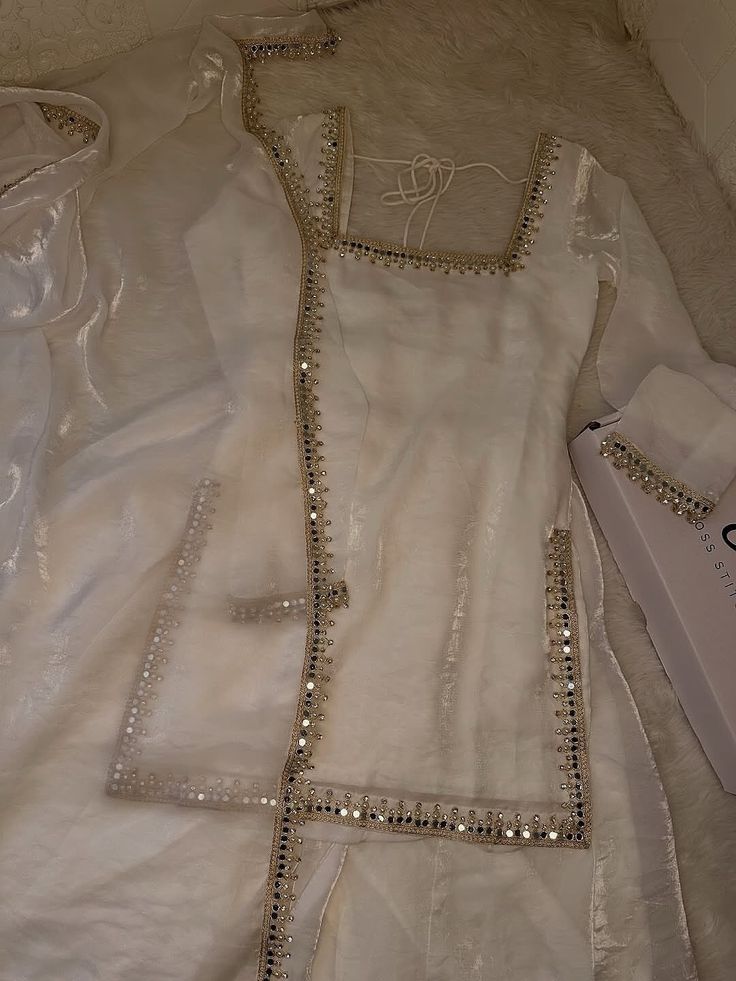 Flat lay of a luxurious white or ivory sheer organza kurta set with heavy silver mirror work (Shisha/Abla) trim along the square neckline, front opening, and hemline. The ensemble includes a matching dupatta and sleeve cuffs, laid out on a soft white surface.